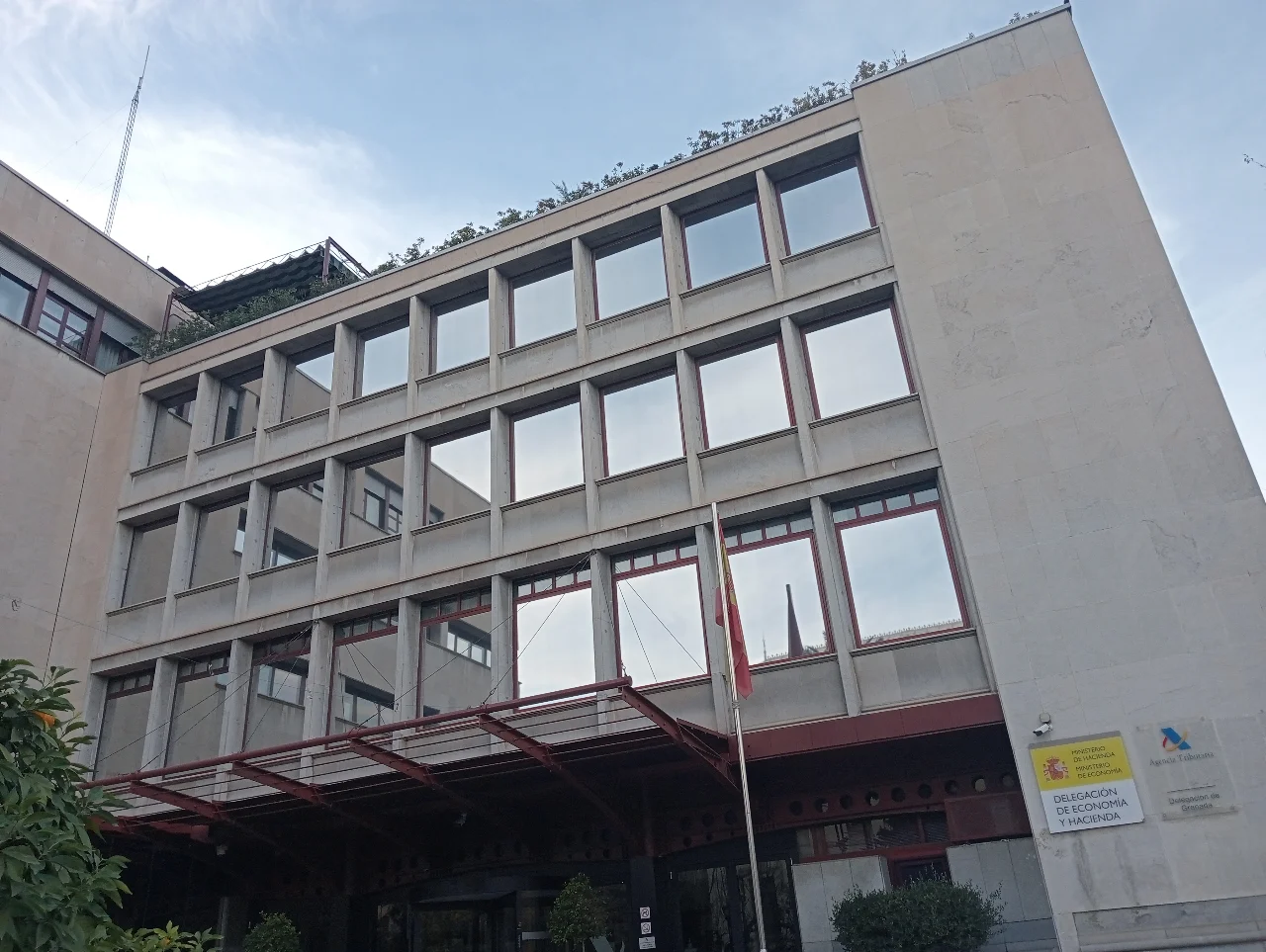 Vue en contre-plongée d’un bâtiment institutionnel avec des fenêtres et des drapeaux ; des enseignes sur la façade indiquent l’Agence fiscale et la Délégation de l’Économie et des Finances à Grenade.